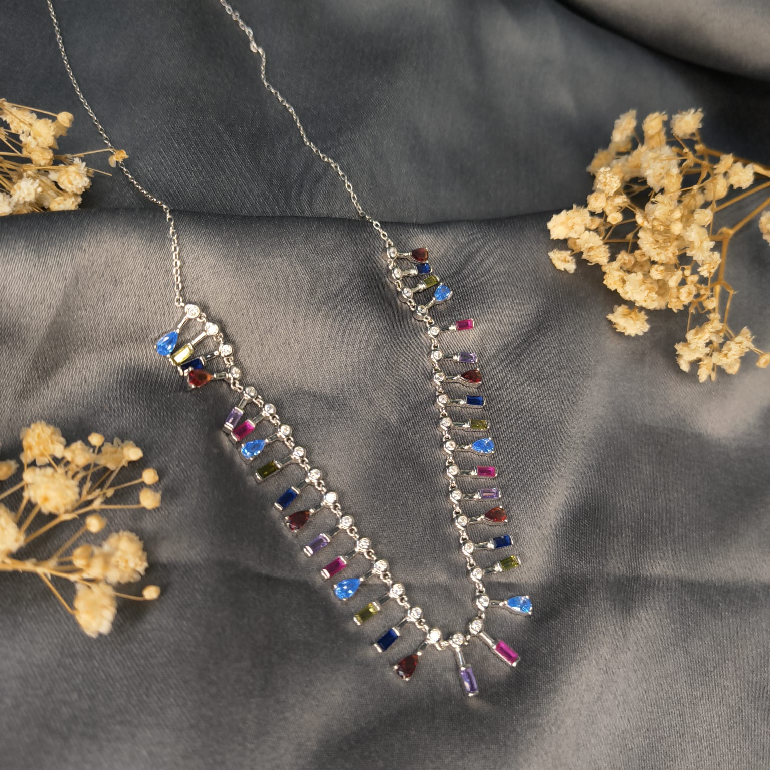 Necklace with multicolored gemstones on a gray fabric background with dried flowers.
