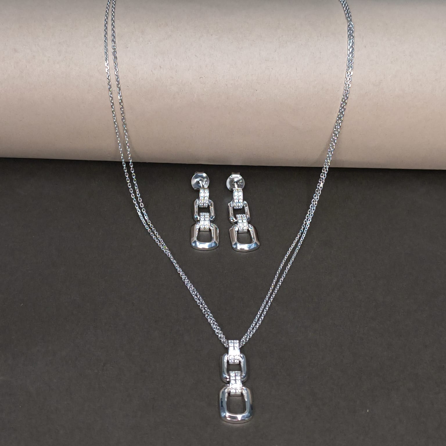 Silver necklace and earrings set on a dark surface with a cardboard roll in the background.