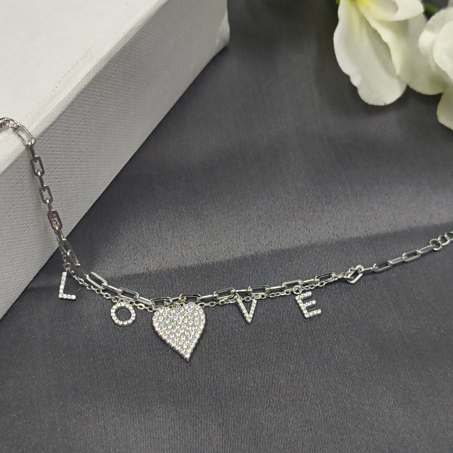 Silver bracelet with heart charm and 'LOVE' letters on a gray surface with white flowers.