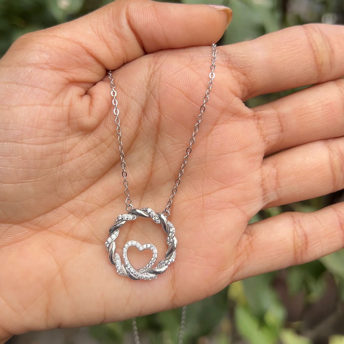 Silver necklace with a heart pendant held in a hand against a blurred green background