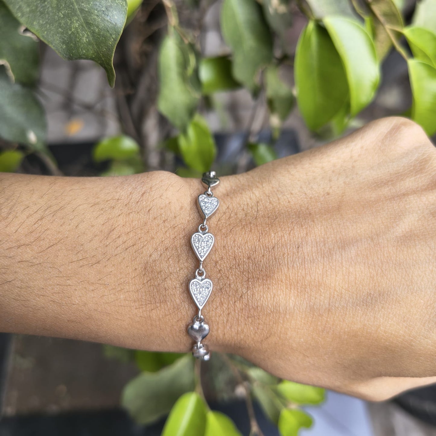 Silver heart-shaped bracelet on a wrist with a blurred green leafy background