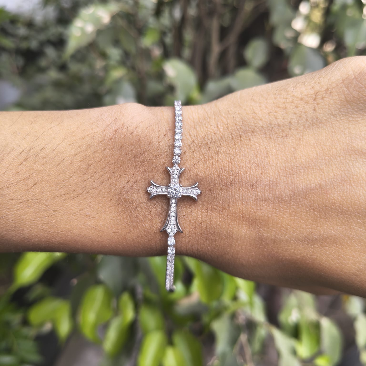 Silver cross bracelet on a wrist with a blurred green leafy background
