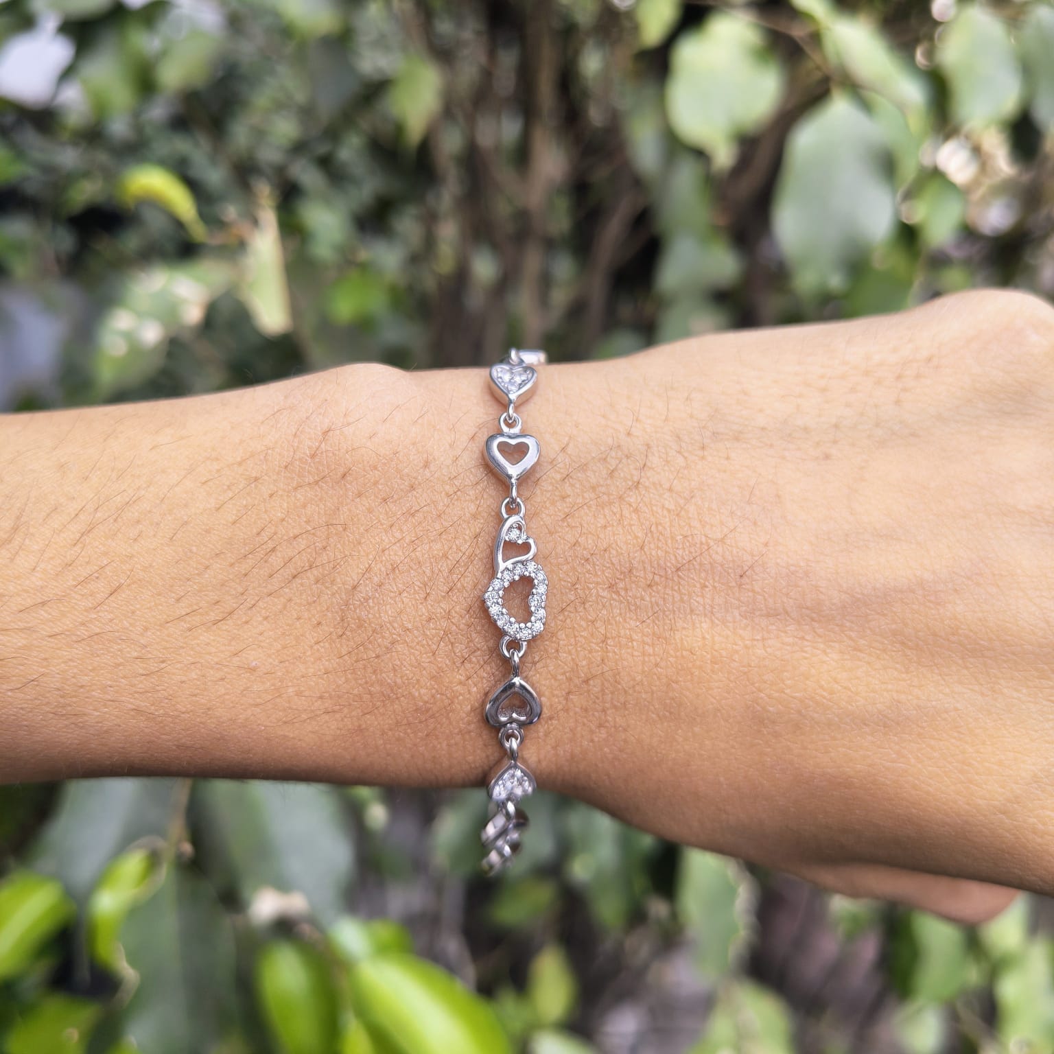 Silver bracelet with heart and circular designs on a wrist against a blurred green background