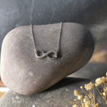 Silver infinity symbol necklace on a dark stone surface with dried flowers.